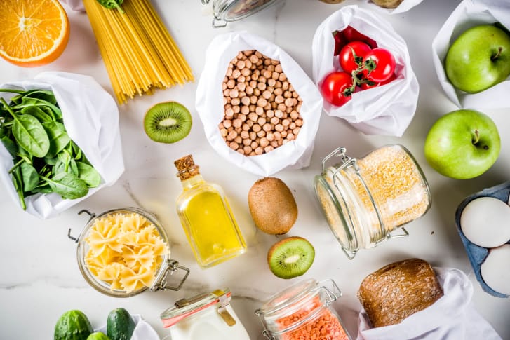 На белой поверхности лежат различные экологичные продукты: фрукты, овощи, крупы, макароны, хлеб.